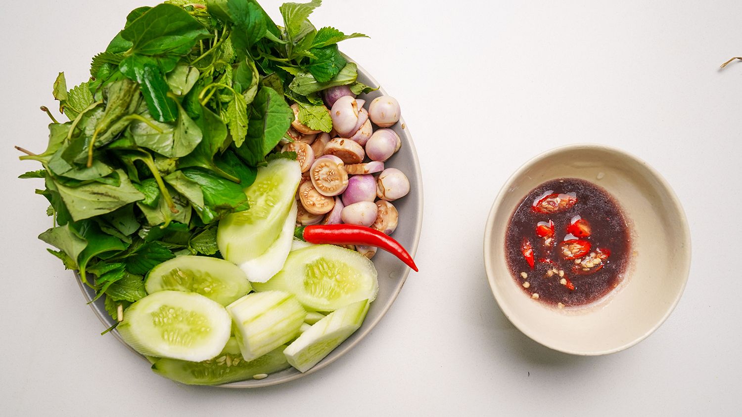 Dĩa rau sống, dưa leo, cà pháo và củ hành kèm chén mắm chấm có ớt tươi trên nền trắng.
