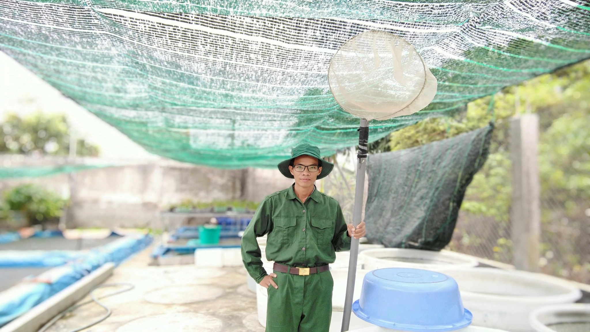 Anh Nguyễn Quốc Đạt mặc trang phục màu xanh cầm vợt lưới cán dài đứng cạnh các bể cá giống
