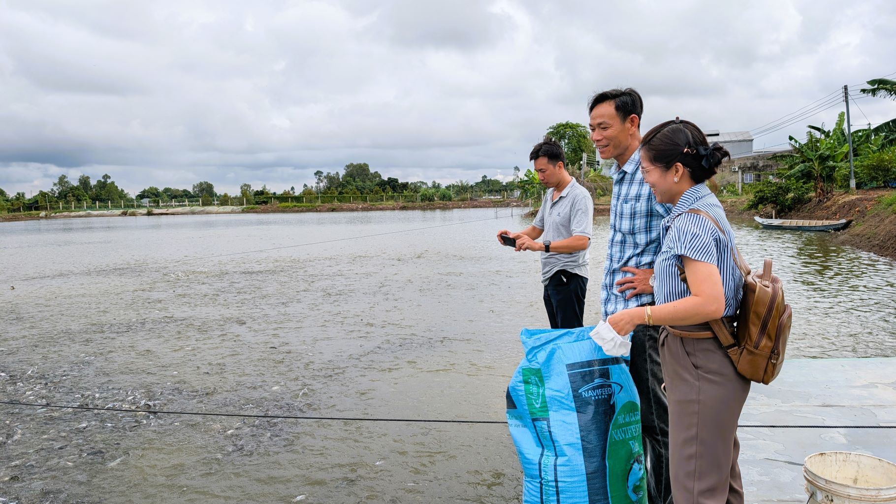 Nhóm người đứng bên ao nuôi thủy sản quan sát cá ăn, bên cạnh có bao thức ăn Navifeed.