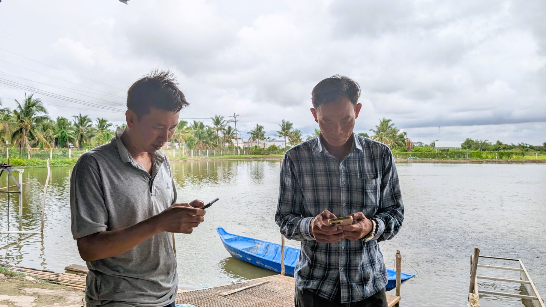 Hai người đàn ông đứng xem điện thoại di động bên cạnh ao nuôi thủy sản.